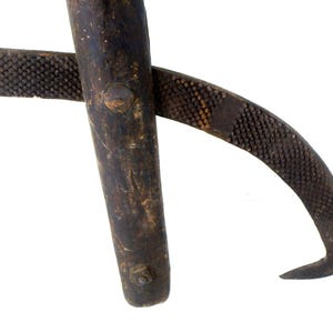 Puede incluir: Primer plano de una herramienta vintage de color marrón oscuro con una hoja curva y texturizada y un mango. El mango tiene un accesorio remachado y una pequeña lengüeta perforada. La herramienta parece ser de metal y muestra signos de desgaste.