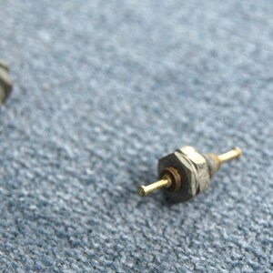 Puede incluir: Primer plano de un componente electrónico plateado con un cable de color dorado. El componente está sobre una superficie azul texturizada.