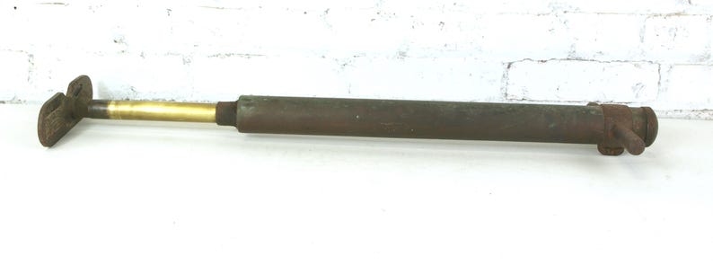 May include: A vintage metal hand pump featuring a long, cylindrical, dark brown rusted body with a smooth, polished brass-colored extension rod and a flat, T-shaped handle at one end, showing signs of age and oxidation throughout the surface.