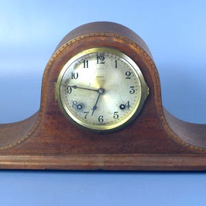 Könnte beinhalten: Eine hölzerne Kaminuhr mit gewölbter Oberseite und rechteckigem Sockel. Das Zifferblatt ist goldfarben mit schwarzen Ziffern und Zeigern. Die Uhr ist aus dunkelbraunem Holz gefertigt.