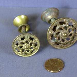 Puede incluir: Dos pomos de armario de latón antiguo con diseños florales ornamentados. Los pomos tienen una base redonda y un vástago con una parte superior decorativa.