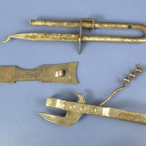 Pode incluir: Uma variedade de ferramentas de cozinha de metal vintage, incluindo um saca-rolhas, um abridor de latas e um abridor de garrafas. As ferramentas são feitas de metal manchado e estão dispostas sobre uma superfície azul. O abridor de latas tem uma alavanca e uma ponta afiada.