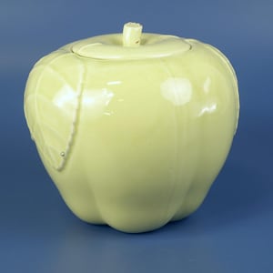 May include: A pale yellow ceramic apple-shaped container with a lid. The container has a stem-like handle on top and a leaf design on the side. The glossy finish reflects light, and the overall design is smooth and rounded.