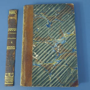 May include: An antique hardcover book with a brown leather spine and a marbled cover featuring diagonal blue and grey patterns. Beside the book lies a detached, worn brown leather spine strip with gold lettering reading Savigny's Obligationen 2.