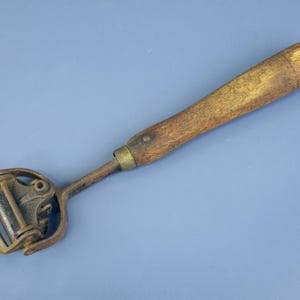 Könnte beinhalten: Ein antiker Käseschneider mit Holzgriff und Metallschneidrad. Der Griff ist hellbraun und die Metallkomponenten weisen Alters- und Gebrauchsspuren auf. Der Schneider ist vor einem blauen Hintergrund platziert.