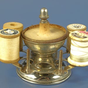 May include: A vintage metal sewing thread spool holder featuring a central circular pincushion with a silver-toned metal cap. The base has multiple vertical pegs for holding wooden spools of thread, with two spools currently attached to the stand.