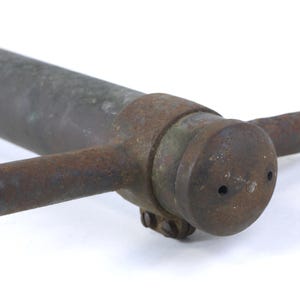 May include: A weathered, dark gray metal pipe featuring a cylindrical end cap with two small circular openings. The end cap is secured by a rusted metal collar that includes two horizontal, rod-like handles extending from opposite sides.