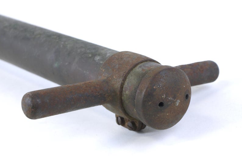 May include: A weathered, dark gray metal pipe featuring a cylindrical end cap with two small circular openings. The end cap is secured by a rusted metal collar that includes two horizontal, rod-like handles extending from opposite sides.