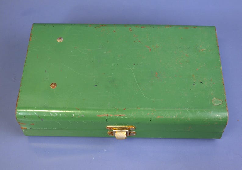 May include: A rectangular, vintage green metal box with a hinged lid and a clasp closure. The box shows signs of wear, with some rust and scratches visible on the surface. The clasp is a gold color.
