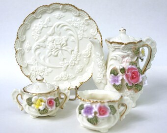 May include: A white porcelain tea set with floral accents. The set includes a teapot, creamer, sugar bowl, and a plate. The teapot has a gold trim and a floral design. The creamer and sugar bowl have a similar design. The plate is plain white with a gold trim.