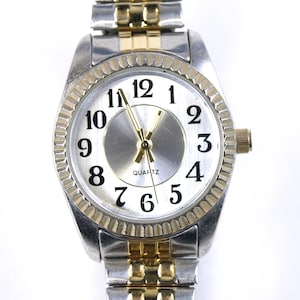 May include: A silver and gold-tone quartz watch with a white face and black numerals. The watch has a fluted bezel and a two-tone metal band. The watch hands are gold-tone.