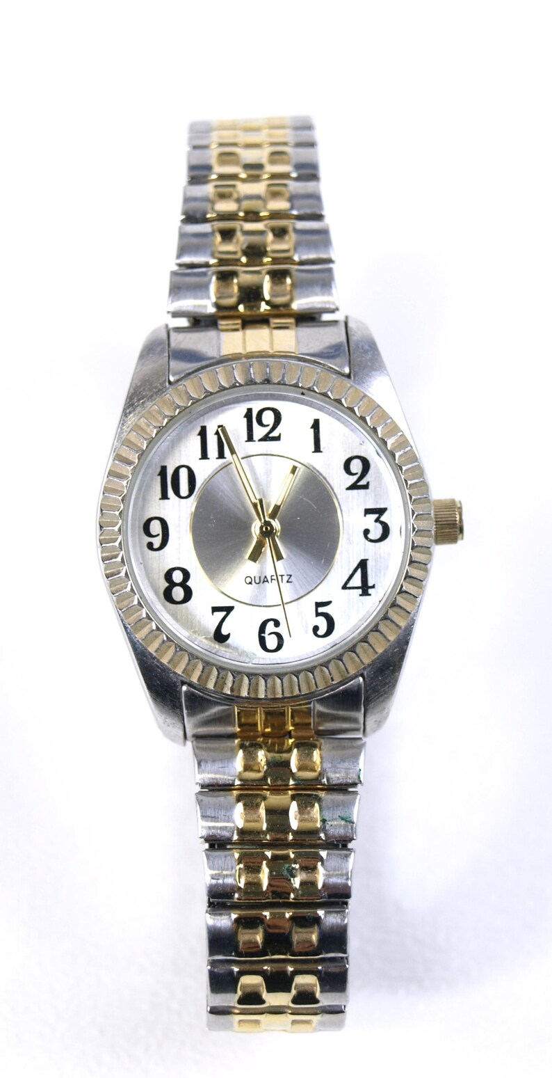 May include: A silver and gold-tone quartz watch with a white face and black numerals. The watch has a fluted bezel and a two-tone metal band. The watch hands are gold-tone.