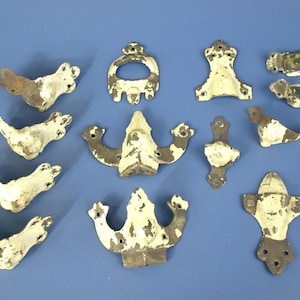May include: A collection of antique metal hardware pieces, including decorative corner brackets, handles, and decorative elements. The pieces are painted white with distressed brown accents, showing signs of age and wear. The hardware is arranged on a blue surface.