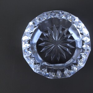 May include: Clear crystal ashtray with a starburst design in the center. The ashtray has a faceted, octagonal rim with decorative, pointed edges. The glass is transparent, reflecting light and creating a sparkling effect. The background is a solid black.