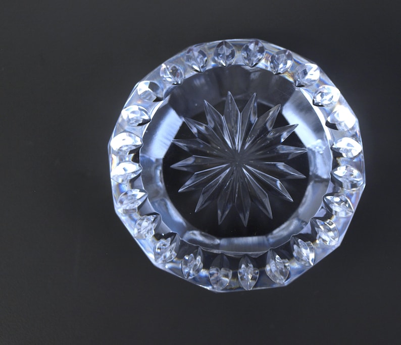 May include: Clear crystal ashtray with a starburst design in the center. The ashtray has a faceted, octagonal rim with decorative, pointed edges. The glass is transparent, reflecting light and creating a sparkling effect. The background is a solid black.