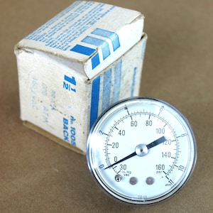 May include: A pressure gauge with a white face, black numbers, and a chrome bezel. The gauge reads up to 160 psi and includes markings for in. Hg vac and atm. A cardboard box with blue stripes and text is in the background.