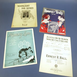 Peut inclure: Quatre partitions de musique anciennes sont présentées sur une surface bleue. Les titres incluent "Moonlight and Roses", "Honest and Truly", "Memory Lane" et "A Little Bit of Heaven". Les couvertures présentent des illustrations et du texte.