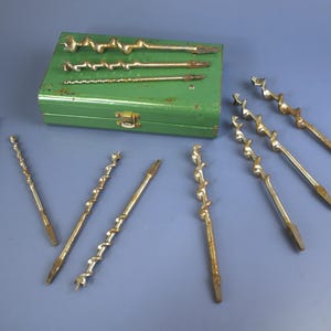 May include: A collection of vintage auger drill bits in various sizes, arranged on a blue surface. The bits are made of metal and show signs of age. A green metal box with a clasp holds additional bits, suggesting a set for woodworking or construction.