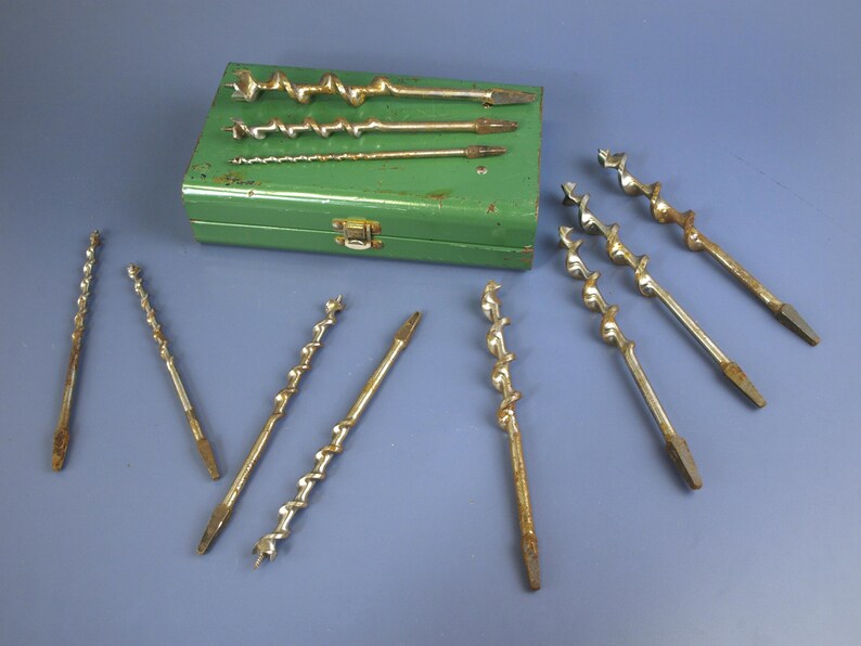 May include: A collection of vintage auger drill bits in various sizes, arranged on a blue surface. The bits are made of metal and show signs of age. A green metal box with a clasp holds additional bits, suggesting a set for woodworking or construction.