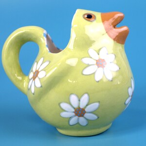 Puede incluir: Jarra de cer&aacute;mica amarilla con forma de pollito, decorada con margaritas blancas. El pollito tiene un pico naranja y ojos marrones. La jarra tiene un asa y un pico. El fondo es azul liso.