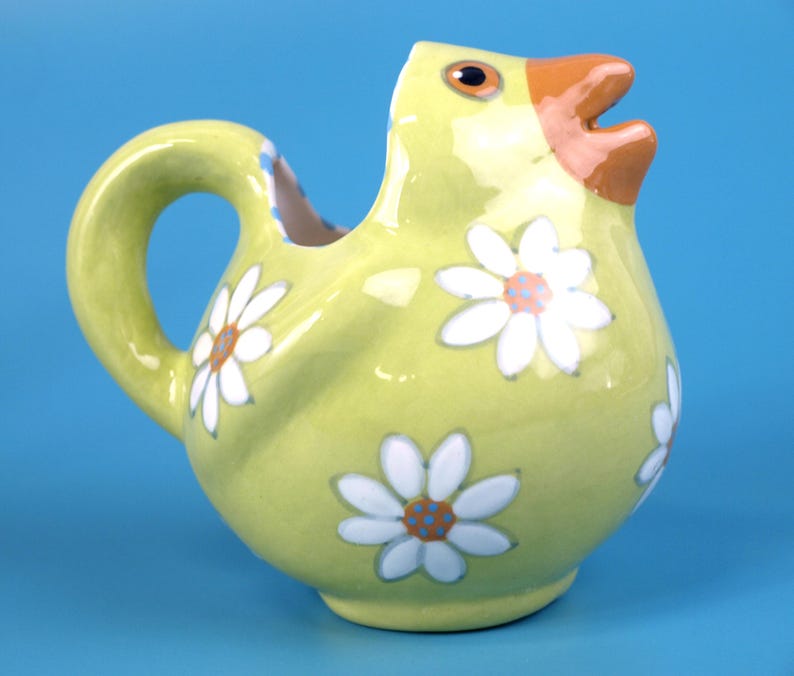 Puede incluir: Jarra de cer&aacute;mica amarilla con forma de pollito, decorada con margaritas blancas. El pollito tiene un pico naranja y ojos marrones. La jarra tiene un asa y un pico. El fondo es azul liso.