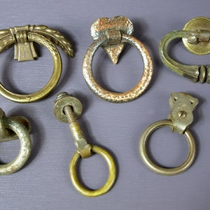 May include: A collection of six antique brass drawer pulls. The designs include a ring with a wheat stalk, a hammered ring with a heart-shaped backplate, and a ring with an ornate backplate. Other pulls include a linked ring, a ring with a bolt, and a simple ring.