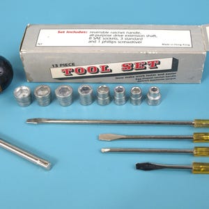 Op de afbeelding: Een 13-delige gereedschapsset op een blauw oppervlak. De set bevat een omkeerbare ratelhandgreep, doppen en schroevendraaiers. De doos leest "TOOL SET" en "Made in Hong Kong."
