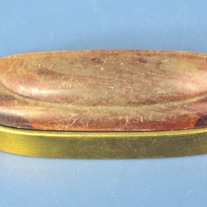 May include: A vintage, oval-shaped box with a brown, textured lid and a gold-colored base. The lid has a raised, curved design. The box is set against a blue background.