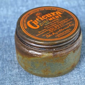 Pode incluir: Uma lata de pomada Cuticura vintage com uma aparência enferrujada e envelhecida. A tampa é marrom escuro com letras laranja e um design circular. O texto na tampa diz "Cuticura Ointment" e outros detalhes. O recipiente é de metal.