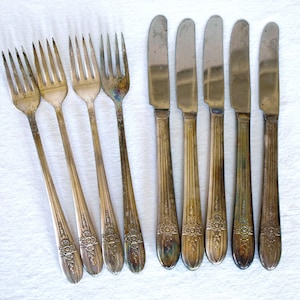 Puede incluir: Un conjunto de cubiertos antiguos de plata, que incluye cuatro tenedores y cinco cuchillos. La cubertería tiene un acabado plateado con detalles florales y lineales ornamentales en los mangos. Las hojas de los cuchillos son redondeadas. La cubertería está dispuesta sobre una superficie blanca.
