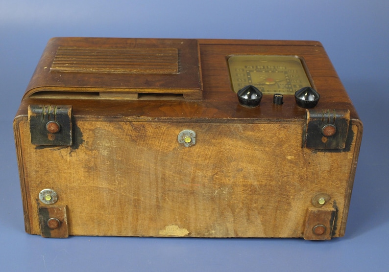 Puede incluir: Una radio vintage de madera con un gabinete de madera marr&oacute;n y una cara negra. La radio tiene dos perillas negras y un dial de vidrio con un fondo dorado.