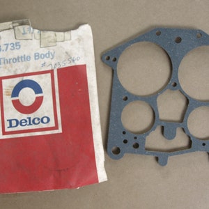 Peut inclure: Un joint en caoutchouc noir pour un corps de papillon. Le joint comporte plusieurs trous et découpes. L'emballage est rouge et blanc avec le texte "Gr. No. 3.735 Gasket Throttle Body #1035540 $1-" et le logo Delco.