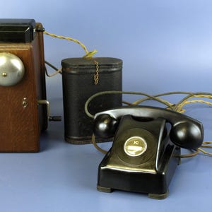 Puede incluir: Un juego de teléfono antiguo con una caja de madera marrón, dos campanas plateadas y un auricular negro. Una batería cilíndrica negra y el cableado completan el dispositivo de comunicación vintage. El teléfono está sobre una superficie azul.