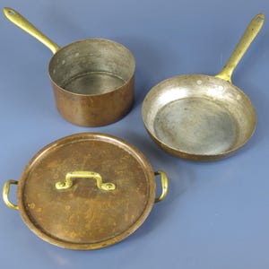May include: Three copper cooking pots with brass handles and a lid. The set includes a saucepan, a frying pan, and a round pot with a lid. The pots have a vintage look and are arranged on a blue surface.