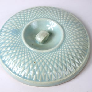 Puede incluir: Una tapa de cerámica azul claro con un patrón de diamantes en relieve y un pequeño pomo en la parte superior.