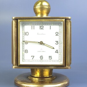 Puede incluir: Un reloj vintage de latón con esfera cuadrada y base redonda. La esfera blanca tiene números y manecillas negras. El texto "Remembrance" está encima del centro, y "SEVEN JEWELS SWISS" debajo. El reloj mide aproximadamente 10 cm de alto.
