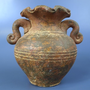 May include: A rustic, weathered brown ceramic vase featuring a wide, ruffled rim and two symmetrical, curled handles on the sides. The body of the vessel is decorated with horizontal incised lines and displays a mottled, textured finish.