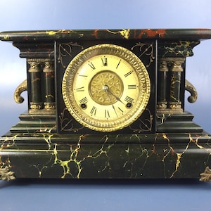 May include: A mantel clock with a black faux marble finish featuring yellow and red veining. The clock has a circular cream-colored face with black Roman numerals, gold-toned decorative columns, and ornate metal feet at each corner.