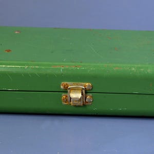 May include: A rectangular, green metal box with a hinged lid and a clasp closure. The box has a slightly worn appearance with some visible scratches and rust spots, suggesting age or use. The clasp is a gold color.