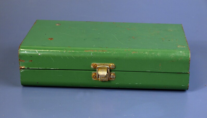 May include: A rectangular, green metal box with a hinged lid and a clasp closure. The box has a slightly worn appearance with some visible scratches and rust spots, suggesting age or use. The clasp is a gold color.