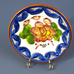 May include: A white ceramic plate with a blue and orange floral design. The plate has a brown rim and a blue and white border. The plate is on a stand.