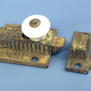 May include: An antique, cast-iron cabinet latch with a white ceramic roller. The latch is a rusty gold color, with a matching strike plate. The hardware is set against a blue background.