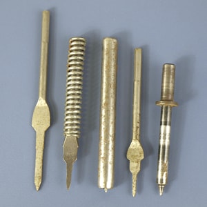 May include: A collection of five metallic industrial components including three pointed needle-like tools with flat sections, one cylindrical rod, and one spring-coiled tool. The items feature varying textures and shades of silver and brass metal.