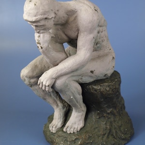 Op de afbeelding: Een verweerde, gebroken witte sculptuur van een zittende figuur in een contemplatieve pose. De figuur rust met de kin op een hand, de andere arm over de benen. De sculptuur is gemonteerd op een getextureerde, donkergroene basis.