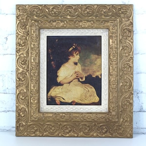 Puede incluir: Una pieza de arte mural rectangular que presenta el retrato de un niño con vestido blanco, dentro de un marco dorado pesado y ornamentado con tallas florales en relieve y un borde interior blanco con un patrón texturizado de puntos.