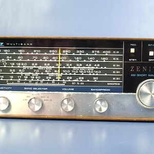 Puede incluir: Radio Zenith AM-Short Wave Receiver vintage con una cara plateada, molduras de madera y múltiples diales y botones. La radio presenta un dial de sintonización con marcas de frecuencia y el logotipo de Zenith.