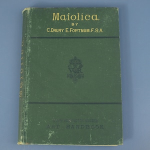 May include: A dark green hardcover book titled Maiolica by C. Drury E. Fortnum F.S.A. The front cover features gold lettering, two horizontal black lines, and a central black emblem. The bottom text reads South Kensington Museum Art Handbook.
