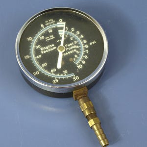 Puede incluir: Un manómetro redondo de vacío de motor y presión de bomba de combustible con esfera negra, marcas blancas y borde de metal plateado. El medidor incluye el texto Engine Vacuum, Fuel Pump Pressure, in Hg vacuum, cm Hg vacuum, kPa y psi con racor.