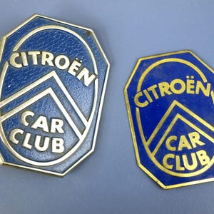 Può includere: Due distintivi vintage del Citroën Car Club. I distintivi sono blu con scritte dorate e un bordo dorato. Il testo "CITROËN" è arcuato in alto e "CAR CLUB" in basso. I distintivi sono ottagonali.