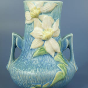 Puede incluir: Un jarrón de cerámica azul con una superficie texturizada y dos asas. El jarrón presenta diseños florales en relieve en blanco, amarillo y verde. El jarrón tiene forma cilíndrica con la parte superior ensanchada.